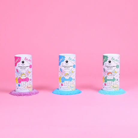 Three colorful bath salt containers stand on a pink background. Each has playful cartoon graphics, and vibrant salts spill out below: purple, blue, and green.