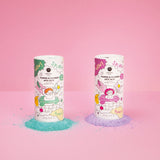 Two colorful, cylindrical containers of foaming bath salts stand on a pink background. Left is turquoise, right is purple, with spilled powder beneath.