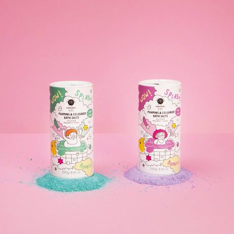 Two colorful, cylindrical containers of foaming bath salts stand on a pink background. Left is turquoise, right is purple, with spilled powder beneath.