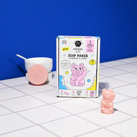 Box of Nailmatic Kids Soap Maker Kit with pink cat illustration on a tiled surface. Nearby, a pink cat-shaped soap and a bowl with a round soap. Playful and colorful.