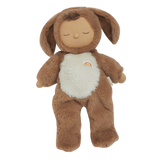 A plush toy with a brown, fluffy dog feature, closed eyes, and a serene expression. It features a white belly with a small embroidered rainbow.