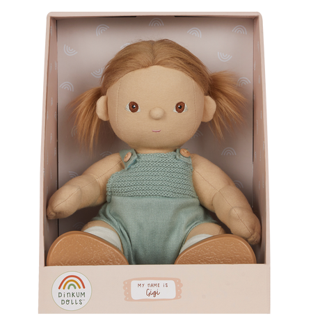 A soft doll with brown hair in pigtails sits in a pastel box. The doll wears a knitted green romper and is labeled "Gigi" with a cheerful expression.