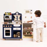A young child opening a white toy refrigerator next to a blue wooden play kitchen set.