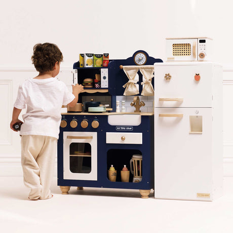 A child playing with a blue wooden toy kitchen set standing beside a white play refrigerator.