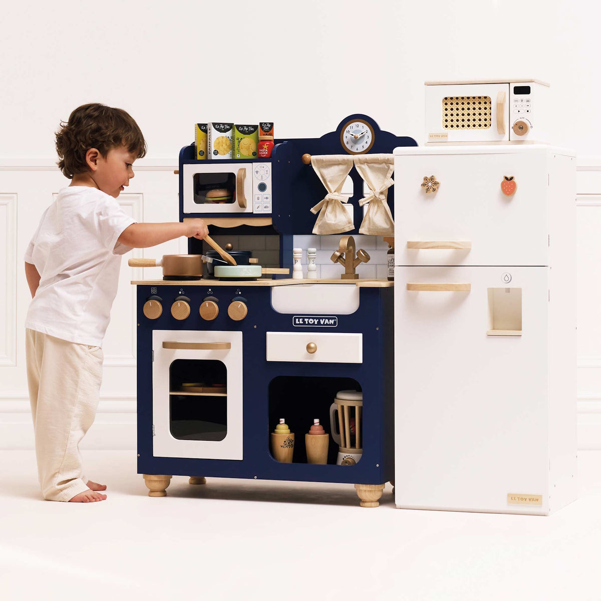 A toddler cooking at a blue wooden toy kitchen set beside a white play refrigerator.