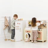Two young children playing with a wooden toy fridge and play kitchen set in a white room.