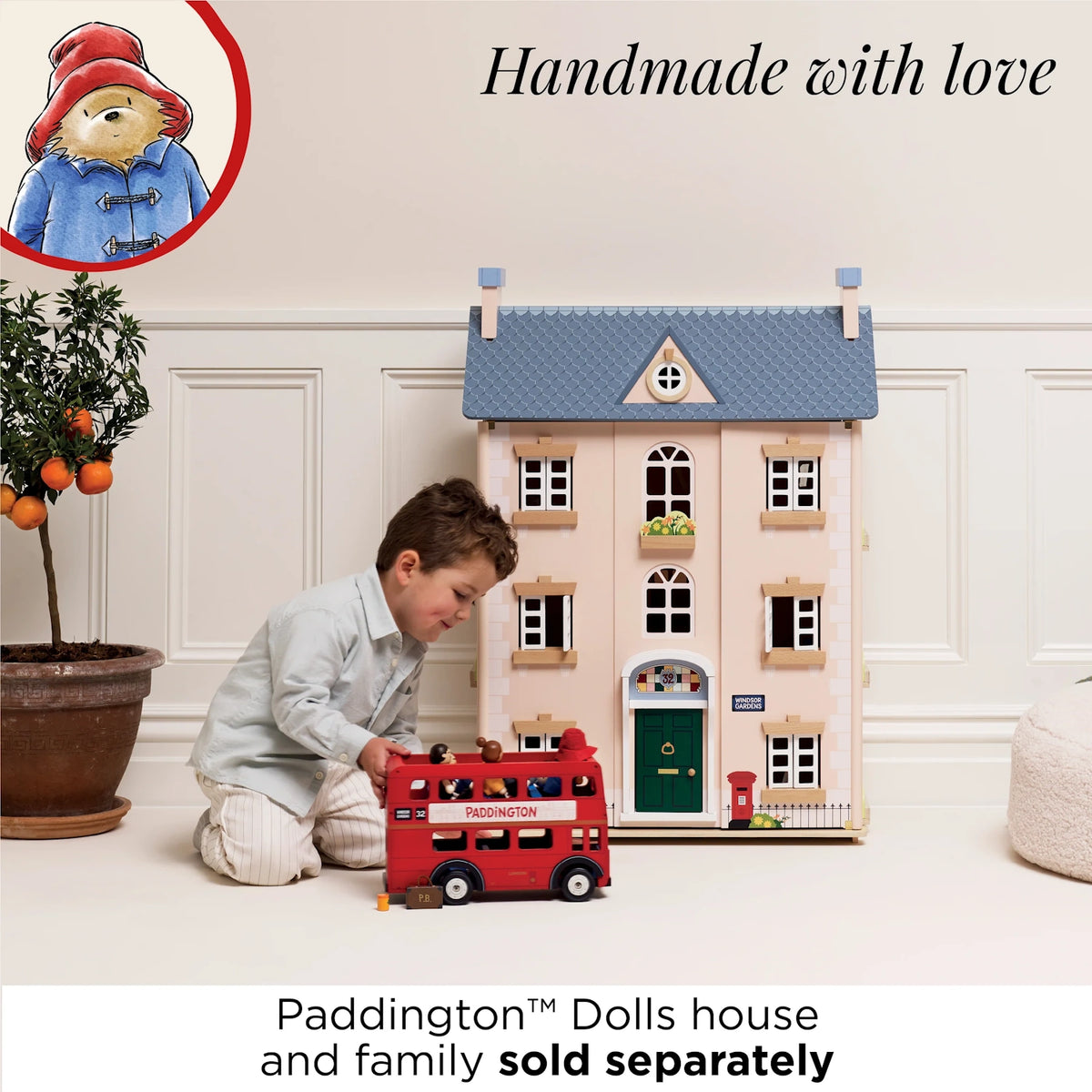 A young boy playing with a red toy London bus in front of a pink wooden dolls house.