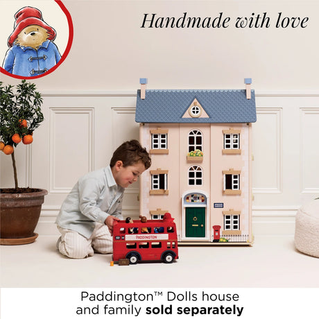 A young boy playing with a red toy London bus in front of a pink wooden dolls house.