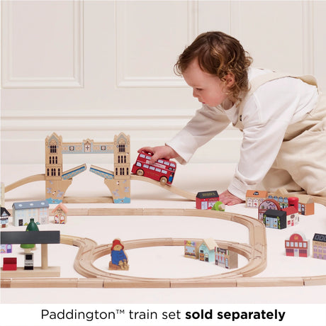 A toddler playing with the Paddington toy bus on a wooden train and bridge track set.