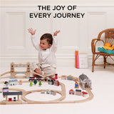 A happy child raising arms while sitting behind an assembled wooden toy train set with Paddington accessories.