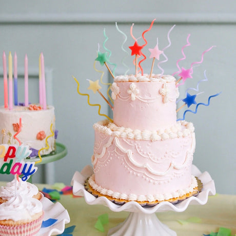 Two-tier pink frosted cake decorated with colorful wavy candles and star toppers on a white scalloped pedestal stand.
