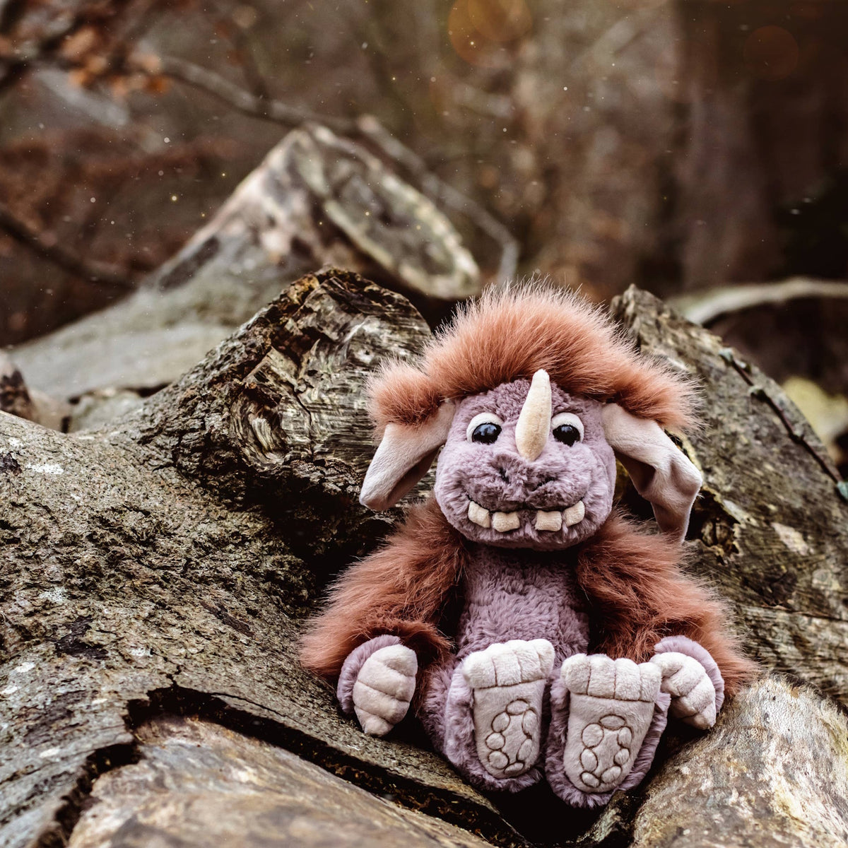 Plush monster toy with grey fur, brown mane, and single horn sitting outdoors on tree logs in a forest setting.
