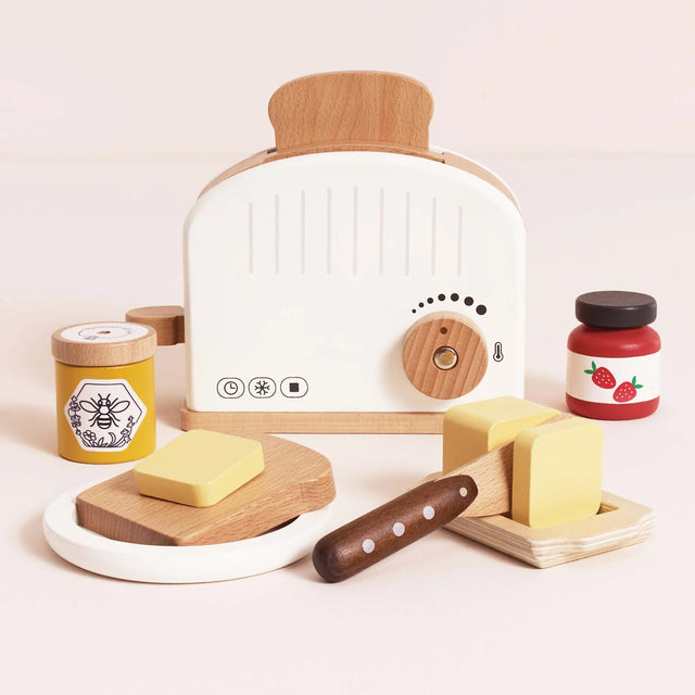 Wooden toy toaster set with a slice of bread, butter, a knife, honey, and strawberry jam on a neutral background.