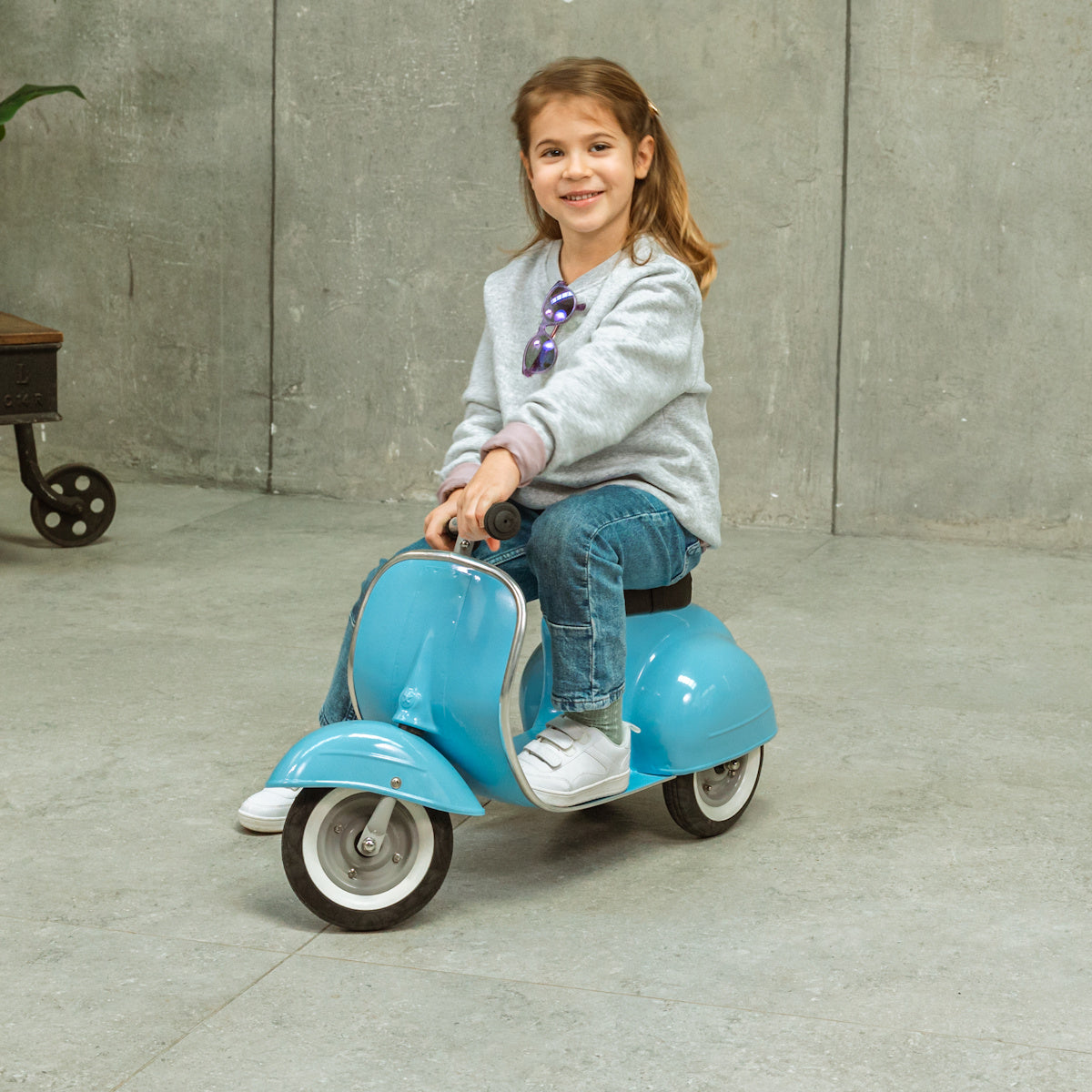 A young girl smiles while sitting on a small blue toy scooter indoors. She wears a grey sweater, jeans, and white shoes, exuding joy and playfulness.