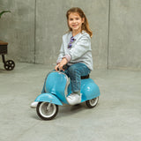 A young girl smiles while sitting on a small blue toy scooter indoors. She wears a grey sweater, jeans, and white shoes, exuding joy and playfulness.