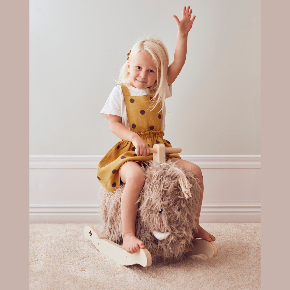 A joyful child in a yellow polka-dot dress rides a fluffy rocking toy, raising one hand in excitement on a soft carpeted floor, exuding playful charm.