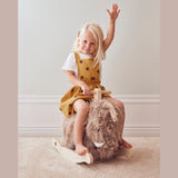 A joyful child in a yellow polka-dot dress rides a fluffy rocking toy, raising one hand in excitement on a soft carpeted floor, exuding playful charm.