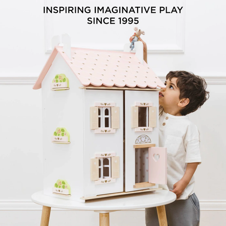 A young child in a white shirt plays with a large, pastel-colored dollhouse on a table. The text above reads, "Inspiring Imaginative Play Since 1995."