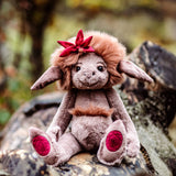 Plush creature with grey fur, brown mane, and red bow sitting outdoors on a wooden log in a natural setting.