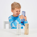 A child in a blue striped shirt is smiling and holding a clear cylinder filled with blue glitter. Two similar glitter cylinders are on a white table.