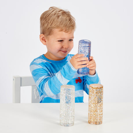 A child in a blue striped shirt is smiling and holding a clear cylinder filled with blue glitter. Two similar glitter cylinders are on a white table.