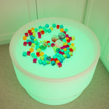 A round table bathed in vibrant green light, scattered with colorful toy blocks and several textured balls. The scene is playful and surreal.