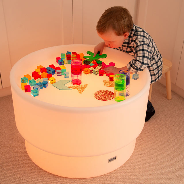 A child in a plaid shirt explores colorful transparent shapes and blocks on a glowing round table, creating a playful and curious mood in a cozy room.