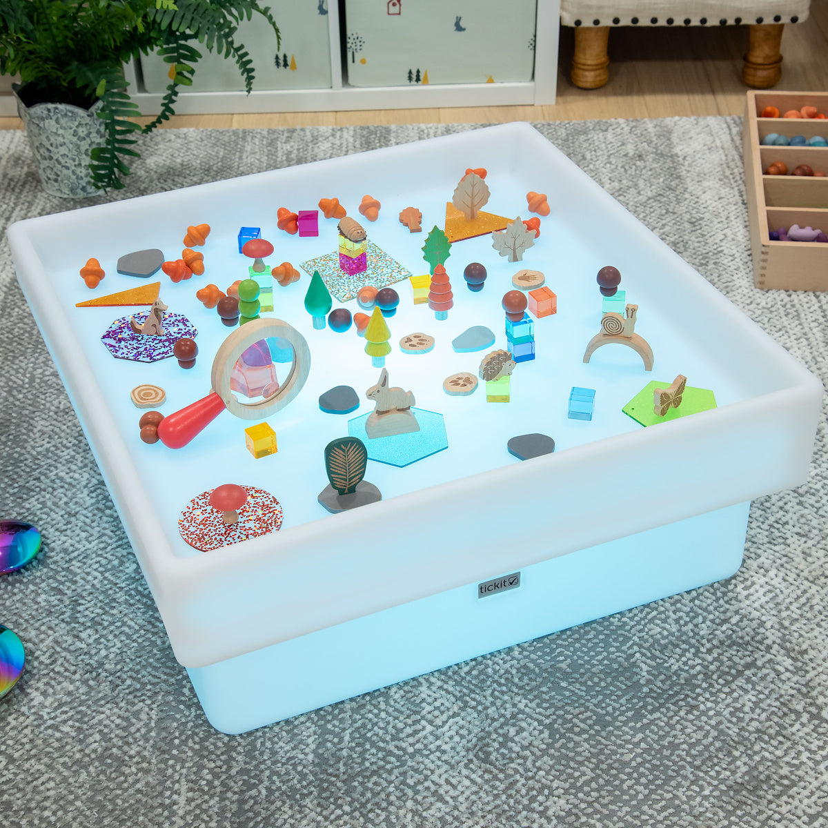 A glowing sensory table with various colorful toys and shapes, including trees, animals, and geometric pieces. The setting is playful and educational.