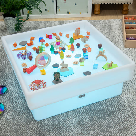 A glowing sensory table with various colorful toys and shapes, including trees, animals, and geometric pieces. The setting is playful and educational.