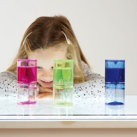 A smiling child with long hair holds a pink liquid timer, surrounded by green and blue timers on a reflective surface. The scene feels playful and colorful.