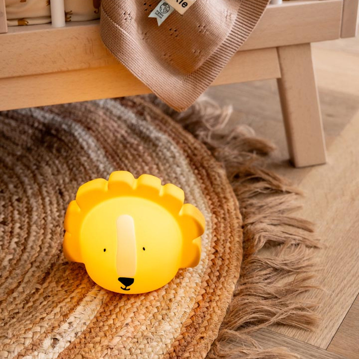 A cute yellow lion-shaped nightlight sits on a woven rug under a wooden crib. A soft beige blanket hangs from the crib, creating a cozy, playful atmosphere.