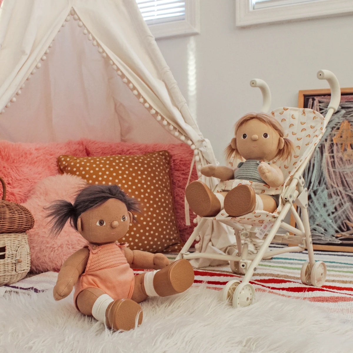 Two plush dolls in a playroom, one sitting on a rug and one in a rainbow-patterned toy stroller.