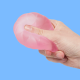 A hand squeezes a pink, flexible, and translucent stress ball shaped like a shrimp against a bright blue background, conveying a playful tone.