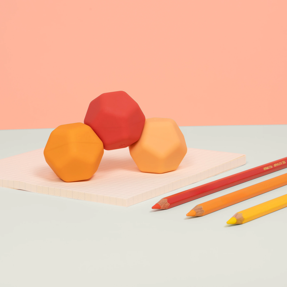 Three faceted balls in red, orange, and peach rest on a grid-patterned pad, with red, orange, and yellow pencils nearby. Background in soft peach tones.