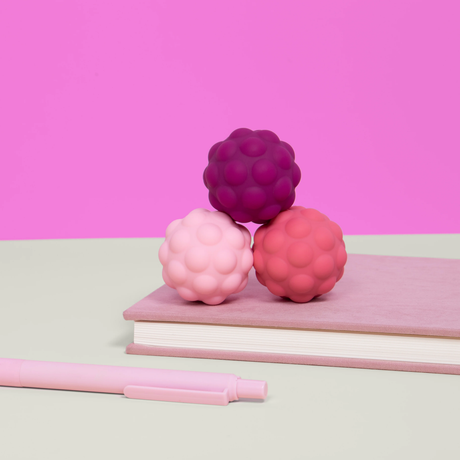 A stack of three textured balls in pink, mauve, and rosy pink sits on a closed book. A pink pen lies beside them. The background is bright pink.