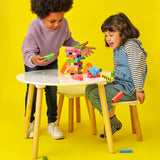 Two children playing a wooden stacking game at a white table, watching colorful blocks fall over against yellow.