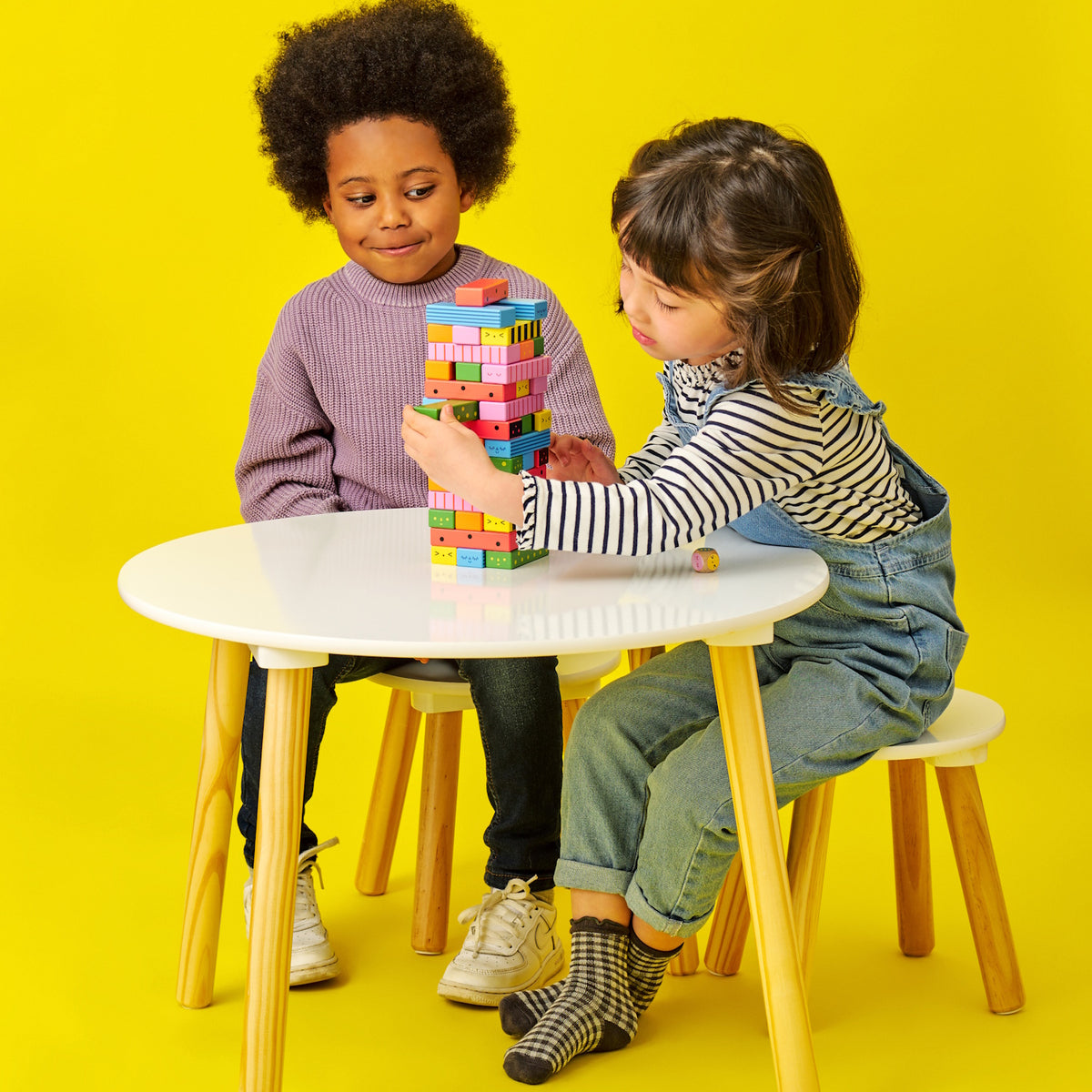 Girl carefully pulling a block from a colorful wooden tower while another child watches at a white table.