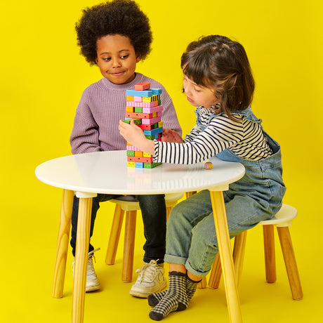 Girl carefully pulling a block from a colorful wooden tower while another child watches at a white table.
