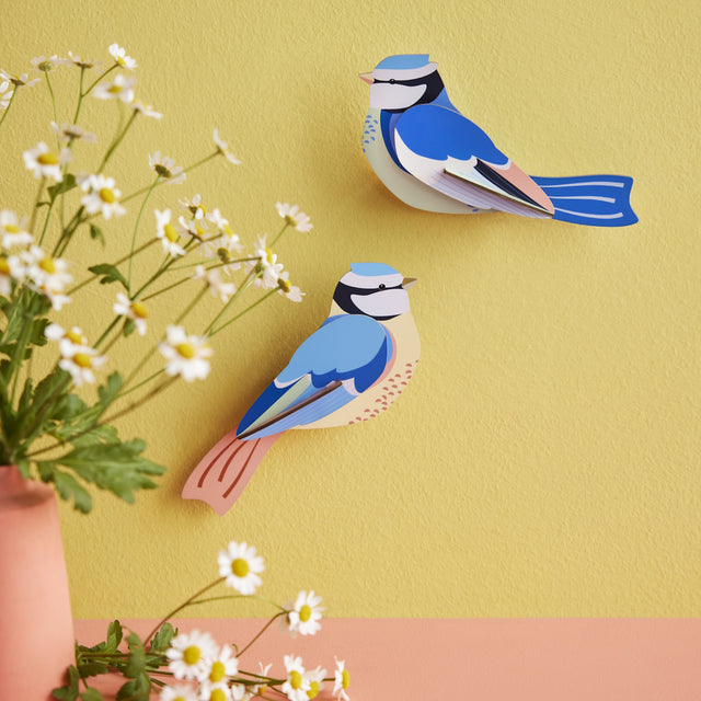 Paper art birds with blue, white, and peach hues on a yellow wall, next to daisies in a pink vase. The scene feels cheerful and artistic.