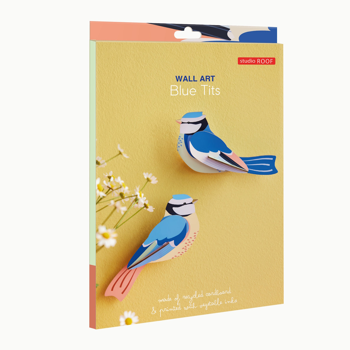 Yellow background showcasing two blue tit birds crafted from layered, colorful cardboard. Text reads "Wall Art Blue Tits" and "made of recycled cardboard & printed with vegetable inks." Flowers accent the bottom left corner.