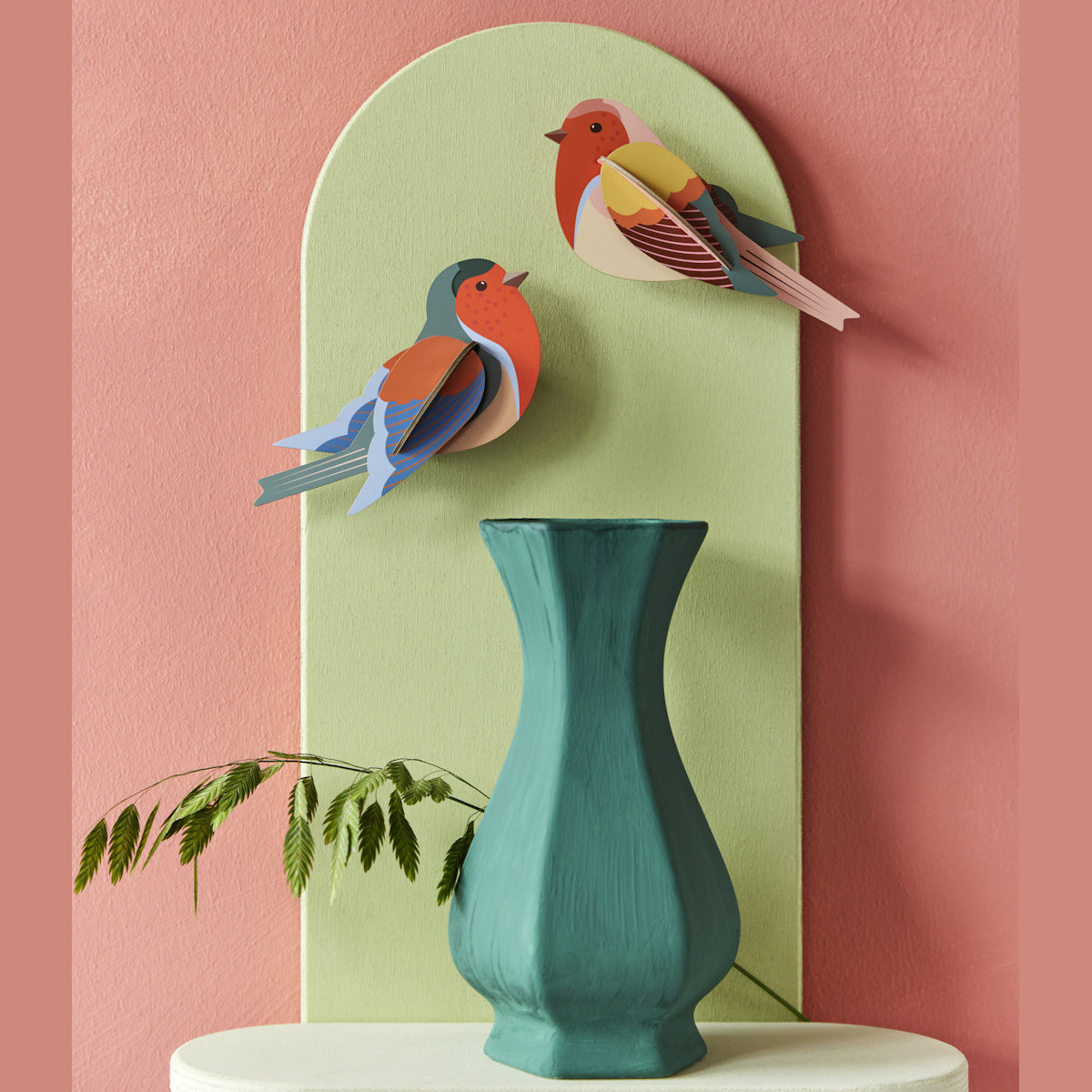 Colorful paper birds are mounted on a pastel green panel against a pink wall. A teal vase with ferns is in the foreground, creating a vibrant and artistic display.