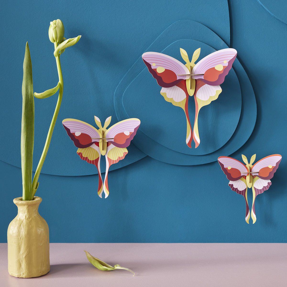Colorful paper butterflies on a textured blue wall create a whimsical scene. A tulip in a yellow vase adds a touch of nature, conveying a serene ambiance.