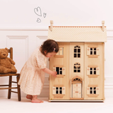 A young child in a cream dress plays with a wooden dollhouse. A teddy bear sits on a chair nearby. The scene is warm and playful, with drawn hearts above.