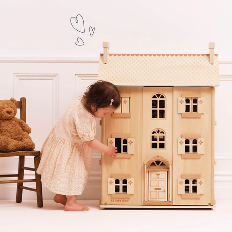 A young child in a cream dress plays with a wooden dollhouse. A teddy bear sits on a chair nearby. The scene is warm and playful, with drawn hearts above.