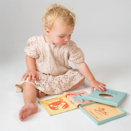 Toddler sitting on the floor reading an open fabric book next to its blue gift box.
