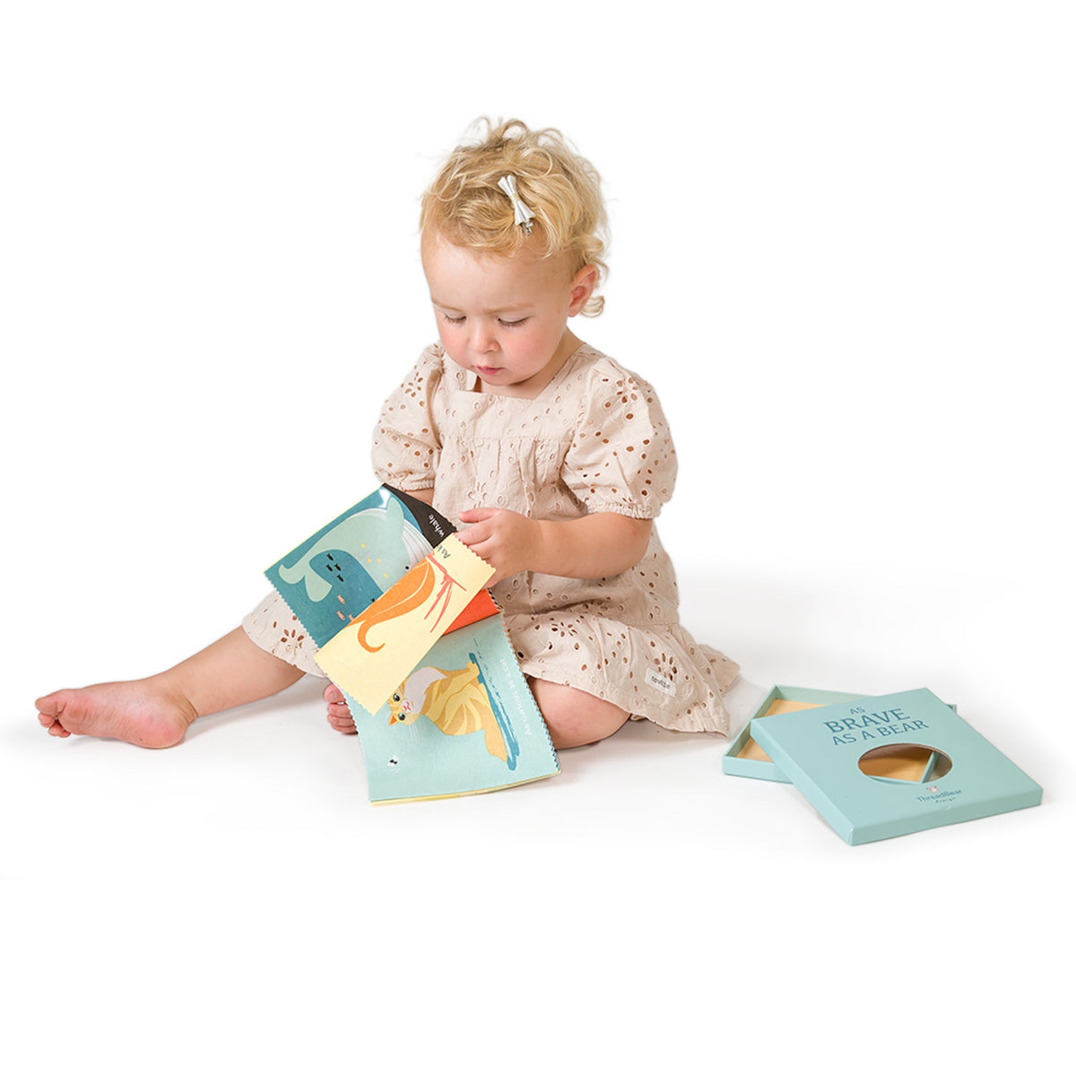 Toddler sitting on the floor flipping through a fabric rag book beside its blue gift box.