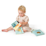 Toddler sitting on the floor flipping through a fabric rag book beside its blue gift box.