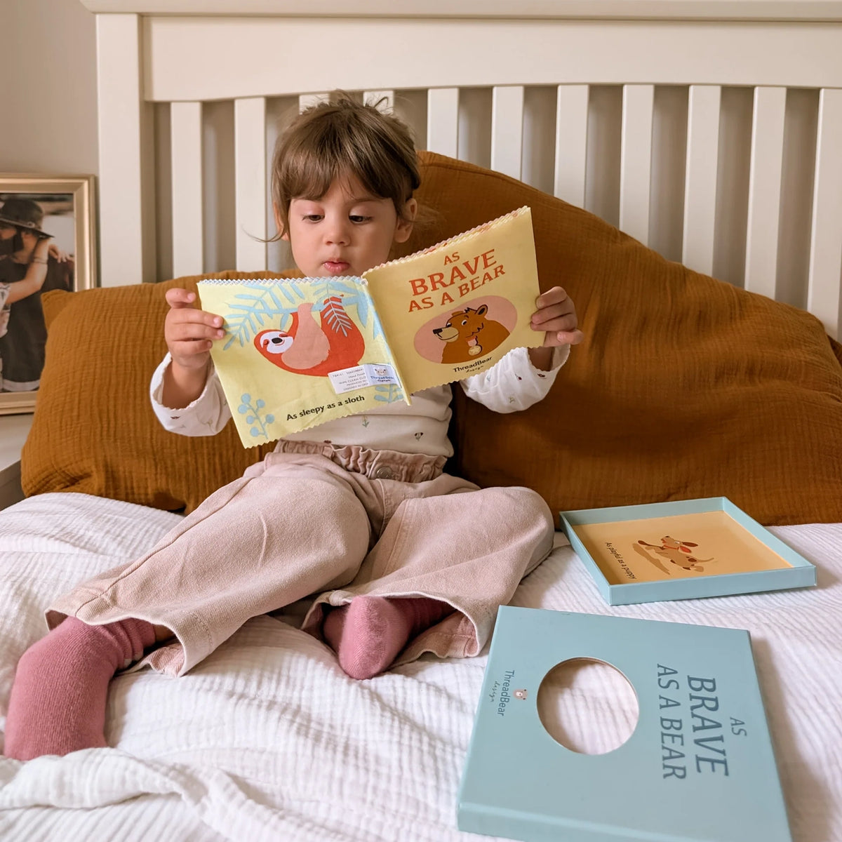 Child sitting on a bed reading an open fabric book titled "As Brave As A Bear" near its packaging.