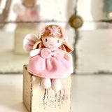 Small plush fairy doll wearing a pink bow dress perched on a distressed wooden block.
