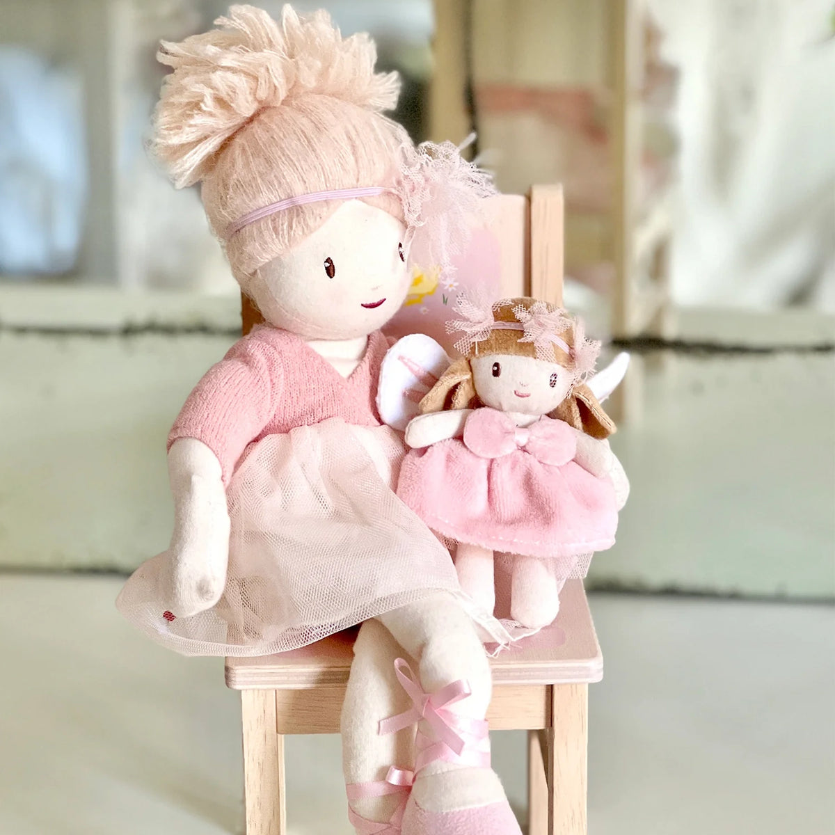 Large rag doll in a pink tutu sitting on a wooden chair holding a small plush fairy doll.